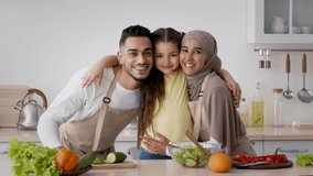 Happy Muslim Family With Kid Girl Hugging Posing In Kitchen. Portrait Of Cheerful Islamic Parents And Little Daughter Standing Together Cooking Dinner At Home. Slow Motion