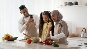 Muslim Family Cooking Dinner Together, Middle-Eastern Husband Taking Photos On Mobile Phone Having Fun Spending Time With Wife And Daughter In Modern Kitchen At Home. Weekend Leisure