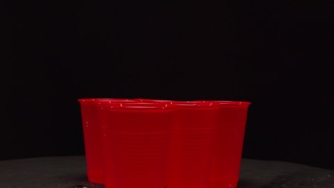 Small orange ball jumps on red plastic cups with alcohol drinks playing beerpong on table in dark studio extreme close view