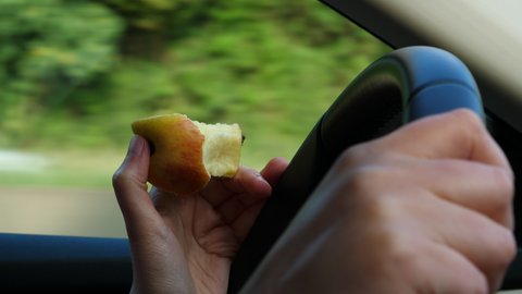 Side view of woman driving fast on highway autobahn holding the leather steering wheel and eating a delicious red apple - Eating or drinking while driving increases the chances of getting car accident