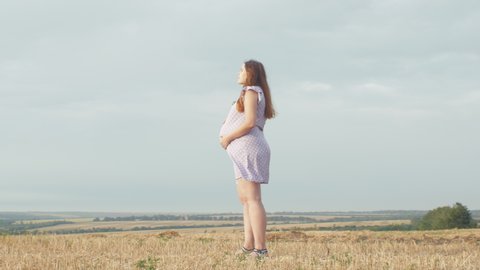camera rotating around happy young pregnant woman in dress standing alone in field on background of cloudy rainy sky, relaxation on nature