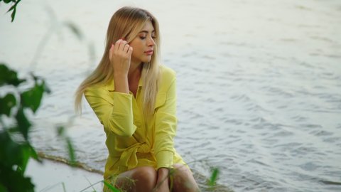  Beautiful blond romantic woman enjoying summer vacation at summer beach. Calm model in yellow outfit hair fluttering in the wind. Portrait of gorgeous girl sitting by water. Travel vacation 4K