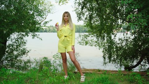 Beautiful blond sexy smiling woman enjoying summer vacation by pond. Model in yellow outfit holding slice of yellow watermelon. Portrait of gorgeous girl eating fresh food 