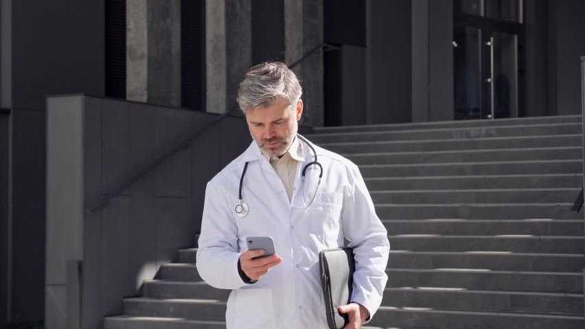 Adult male doctor walking holding laptop texting message, scrolling screen of mobile phone. Middle age doctor sends an online prescription to the patient during a pandemic standing outside clinic. Royalty-Free Stock Footage #1080251063