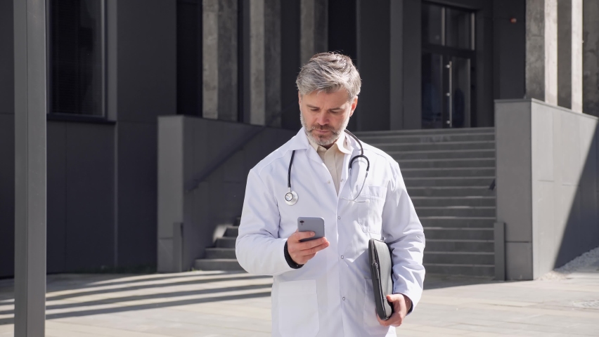 Adult male doctor walking holding laptop texting message, scrolling screen of mobile phone. Middle age doctor sends an online prescription to the patient during a pandemic standing outside clinic. | Shutterstock HD Video #1080251063