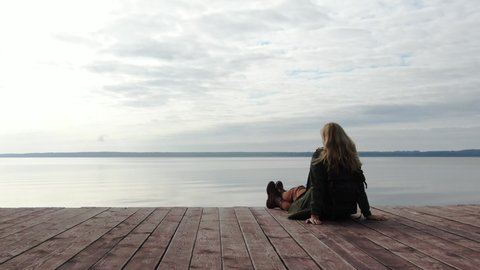 Young woman is sitting on a wooden pier. Enjoys the sea view. Beautiful panorama. Female silhouette at sunset. Meditation. In harmony with nature. Peace of mind and calmness. Traveler on vacation.
