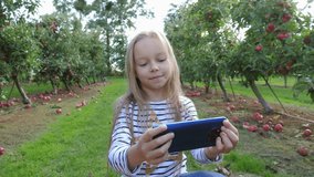 Little cute girl on nature apple tree garden sit watch smartphone while taking selfie grimace shows tongue