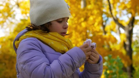 A sick child blows his nose into a handkerchief in the park with a runny nose. Portrait of a girl in the autumn park.