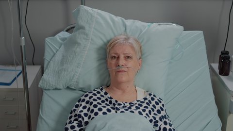 Portrait of old patient with disease having nasal oxygen tube while laying in bed. Close up of senior woman looking at camera and receiving healthcare treatment in hospital ward.
