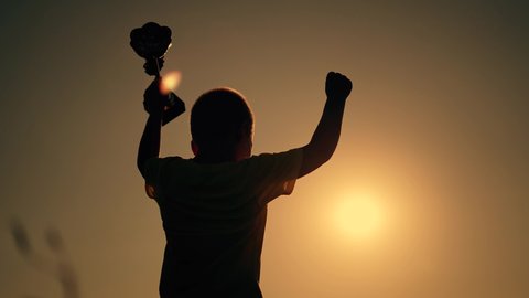 Winner child silhouette. Kid with cup of success raises her hand. Silhouette of child at sunset. Winner success. Victory, path to dream. Child won the victory. Cup in hand of a kid. Superhero kid