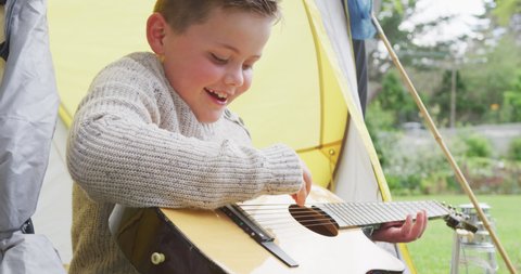 Happy caucasian boy sitting in tent in garden and playing guitar. spending time alone at home.