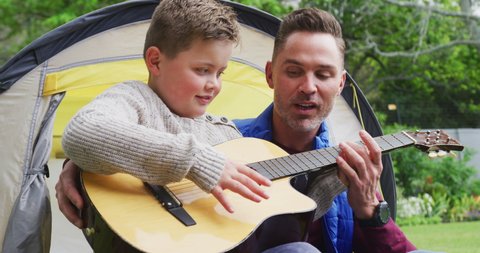 Happy caucasian father with son sitting in tent and playing guitar in garden. family spending time at home.
