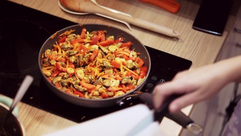 Women's hands pours garlic from cutting board into frying pan with seafood and vegetables and mixes all ingredients with wooden spoon. Dish of shrimp, mussels, carrots and bell pepper.