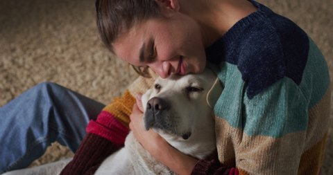 Cinematic authentic close up shot of young happy smiling woman is caressing and kissing with affection her lovely Labrador Retriever dog while having fun together on carpet in living room at home.