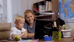 A smiling adult mother and her young son are holding a video conference with a teacher during distance learning, they are sitting at a desk at home against the background of bookshelves.