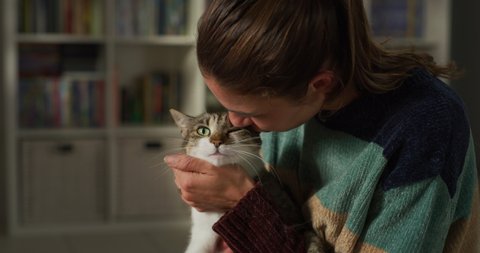 Cinematic authentic close up shot of young happy smiling woman is caressing and kissing with affection her lovely pet cat while having fun together on carpet in living room at home.
