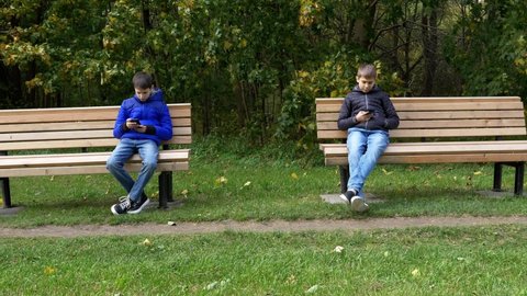 Teen cell phone, video game, social media addiction. Teenage boys sitting on wooden park bench apart and using mobile cell phone instead of communicating, talking or playing together outdoors