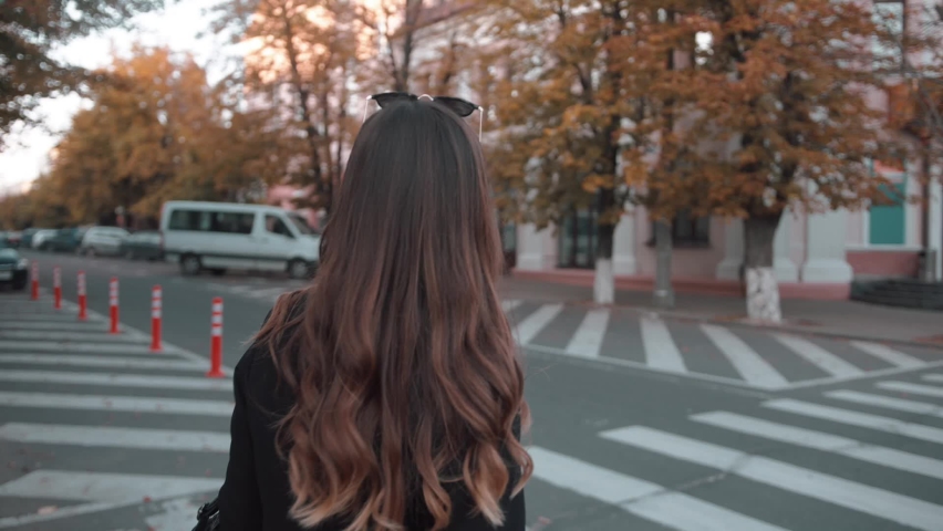 An elegant young girl in a stylish black outfit crosses the road at a pedestrian crossing and straightens her gorgeous hair