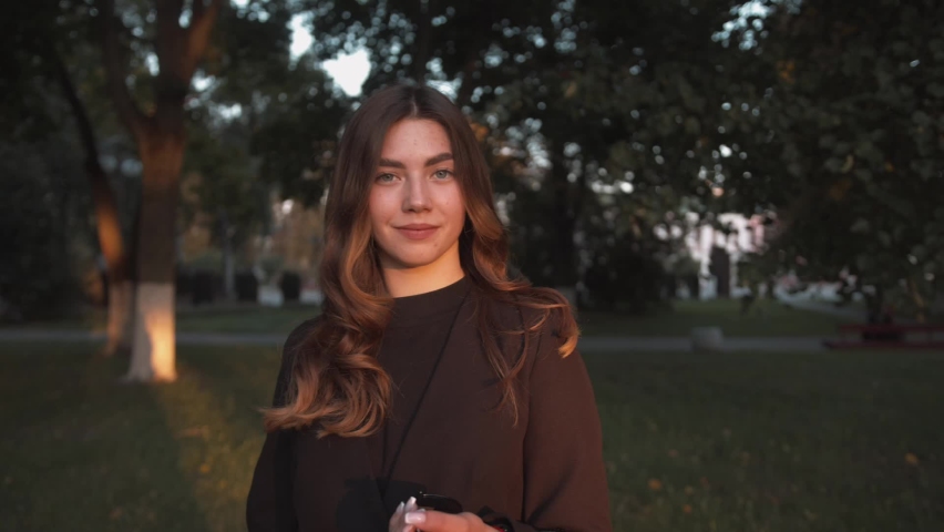 Portrait of a beautiful young girl in black stylish clothes standing among the trees on a city street and laughing. Slow motion