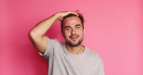 Hey you! Lovely coquet blond man, winking playfully, flirting, smiling, laughing with teeth. Male wear grey t-shirt and fix his hair isolated on pink background. 