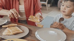 Handheld medium close-up with slowmo of caring young Asian woman spreading honey on bread making toast to her cute little son having breakfast together at home - Powered by Shutterstock - Get 15% off with code: PIKWIZARD15