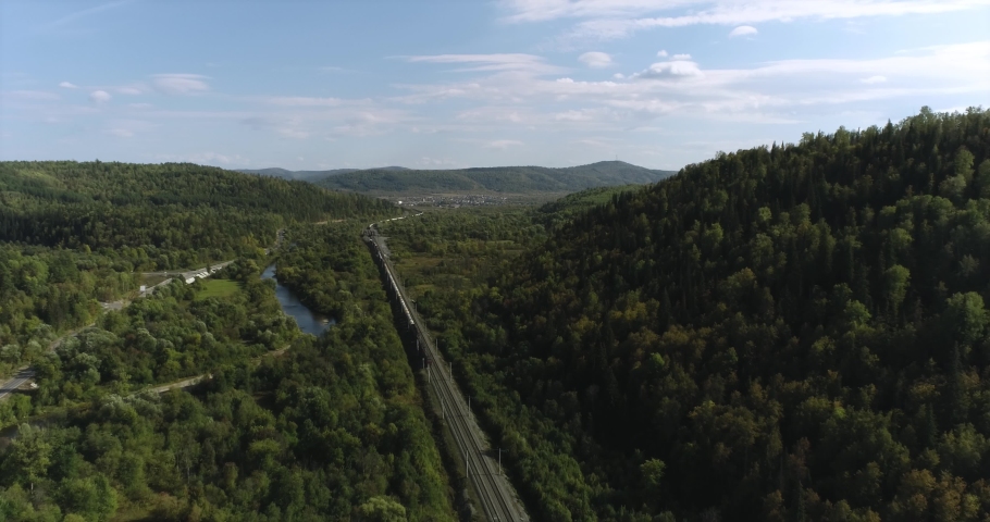 Freight long train carries with oil tank and petrol carriages an electric locomotive by Trans Siberian railways under the rock and near mountain river. Aerial drone wide view at summer sunny day Royalty-Free Stock Footage #1080276704