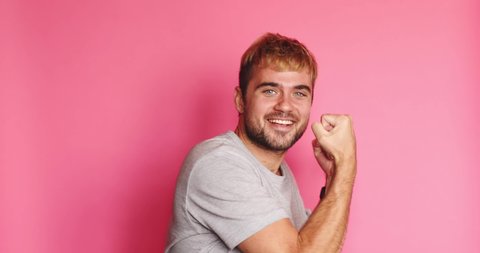 Handsome jubilant overjoyed blond beard man wears grey t-shirt, smart watch looks intently, doing winner gesture celebrate clenching fists say yes isolated on pink background. 