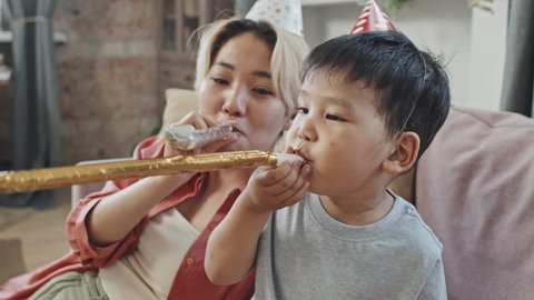 Medium close-up with slowmo of cheerful Asian mother and little son wearing funny Birthday hats blowing party horns at camera celebrating Birthday at home