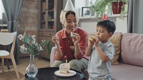 Medium slowmo shot of Asian mother and 3-year-old son in Birthday hats blowing noisemakers celebrating Birthday together at home