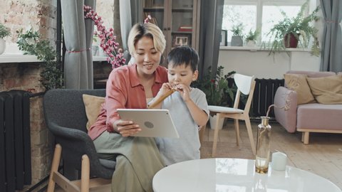 Medium slowmo shot of happy young Asian woman and her cute 3-year-old son having video chat with family or friends on digital tablet at cozy apartment. Boy blowing party horn