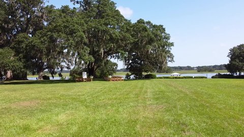 Top view of the wedding venue in a green field under the tree. Tradition of wedding ceremony with decorated decor on the lawn. Rows of festive chairs and beautiful flowers in baskets. Lake shore