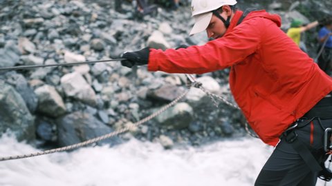 A man crossing a fast mountain river during a hike. A tourist crosses the river using vertical ropes, fastened with a carabiner. Side view. A tourist crosses a fast mountain  river