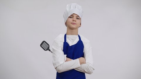 Confident little chef posing with slotted turner at white background. Portrait of Caucasian boy in cook hat and apron crossing hands looking at camera with serious facial expression