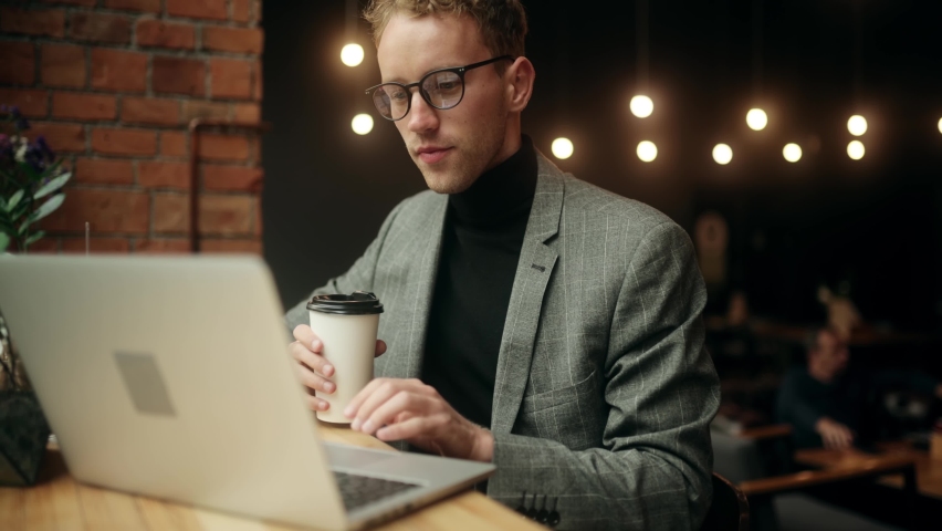 Successful young businessman using laptop while sitting in cafe. Attractive young man with cup of coffee texting on laptop. | Shutterstock HD Video #1080316409
