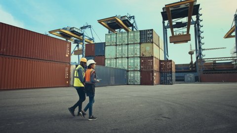Multiethnic Female Industrial Engineer with Tablet and Black African American Male Supervisor in Hard Hats Walk in Container Terminal. VFX Double Girder Gantry Cranes Work in the Background.