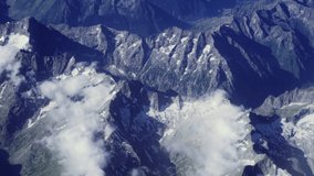Travel flying above snow caped mountain range, looking to the right of the frame. Passenger unobstructed window view of the Alps with clouds, aboard an inclining airplane cruising at high altitude.