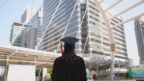 Back view of beautiful young woman in graduation gowns standing smiling holding diploma and hands raise up on outdoor in city. Happy asian newly graduated female successful looks to future and proud. 