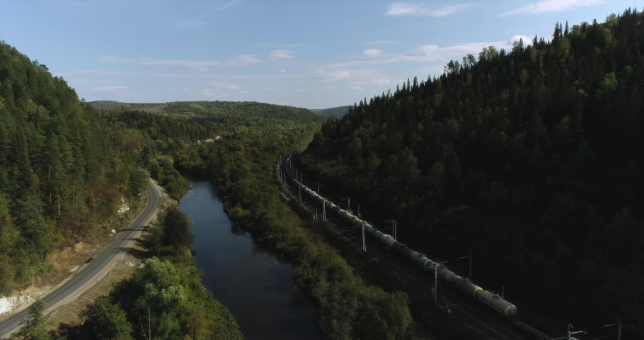Freight long train carries with oil tank and petrol carriages an electric locomotive by Trans Siberian railways under the rock and near mountain river. Aerial drone wide view at summer sunny day Royalty-Free Stock Footage #1080333488