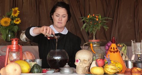Woman witch make potion in glass bowl with Halloween decorations and skull on table. Goth lady preparing potion and pouring poison from bottle. Halloween potion making. Starts from blurred background