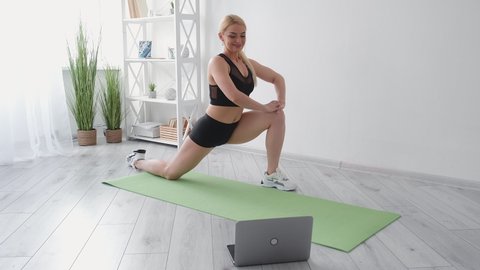 Online fitness. Sportive woman. Home gym. Healthy lifestyle. Happy athletic lady in black sportswear doing stretching workout legs split on yoga mat looking laptop in light room interior.