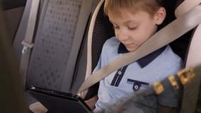 Close-up of a little boy sitting in a child car seat with seat belts and playing video games on a tablet.