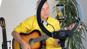 charming elderly man in a yellow shirt plays guitar in front of a ring light and a smartphone, teaches students online skills, records a training video, concept performance, Home Recording Studio