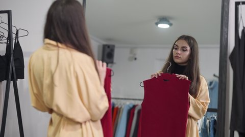 Reflection in shop mirror of focused Caucasian beautiful woman trying on red sleeveless shirt thinking. Concentrated buyer choosing clothes in retail store on Black Friday sales season. Fashion style