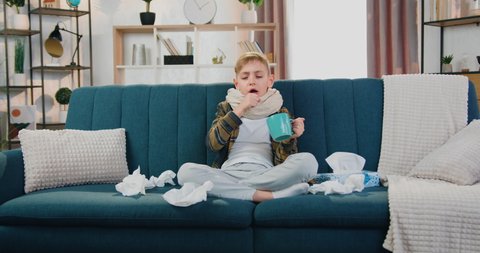 Portrait of cute sad ill teen boy with scarf around neck which sitting on the couch at home surrounded with napkins during influenza,he drinking hot tea and coughing