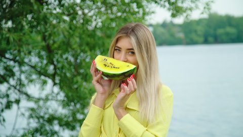 Beautiful blond sexy smiling woman enjoying summer vacation at beach. Model holding yellow watermelon in front of her face. Portrait of gorgeous girl with green eyes looking at camera. She sniffs food