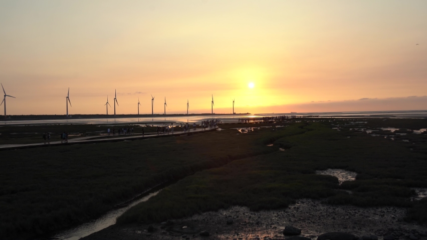 Spectacular orange sunset landscape shot of tourists strolling on wooden walkway platform at beautiful Gaomei wetlands preservation area with wind power plant in the background in Taichung, Taiwan.