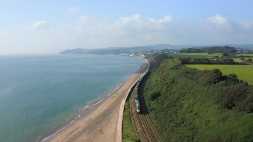 An aerial shot of a long train running along the South Devon coastal railway with the blue sea and seaside views heading towards Dawlish town on a sunny summers day Royalty-Free Stock Footage #1080358382