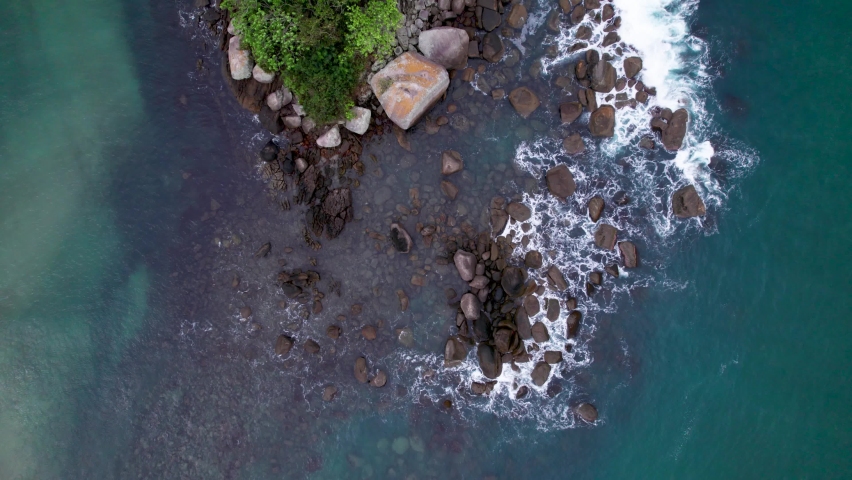 Waves on a small island are seen from above., blue and white water hit the rocky shore. Whitetip waves and dark contrast rocks. Ilhabela, Brazil.