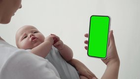 Young Mum with Baby Look Phone with Green Screen Chroma Key Close-up. Mom Use Hold in Hand Kid and Cell Phone Mock-up. Concept Watch and Show Internet Social Media, Calls, Read Blog, App, Iot in Home