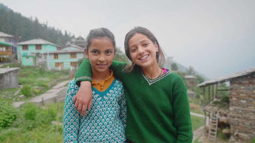 A mid shot of two Indian Asian rural or countryside happy young school girls or kids standing together smiling and looking at the camera with the mountain village in the background